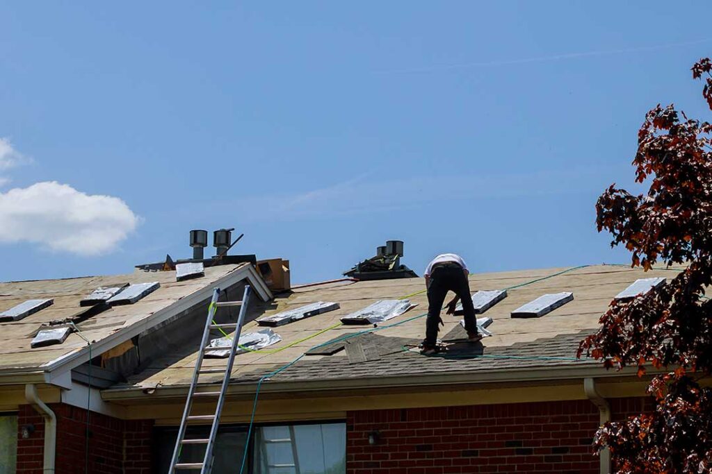 A person stands in a backyard looking up at a house roof covered in debris and damaged shingles after a storm.