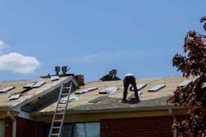 A person stands in a backyard looking up at a house roof covered in debris and damaged shingles after a storm.