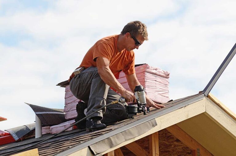 A homeowner examines a ceiling stain beneath a roof leak while water drips into a bucket.